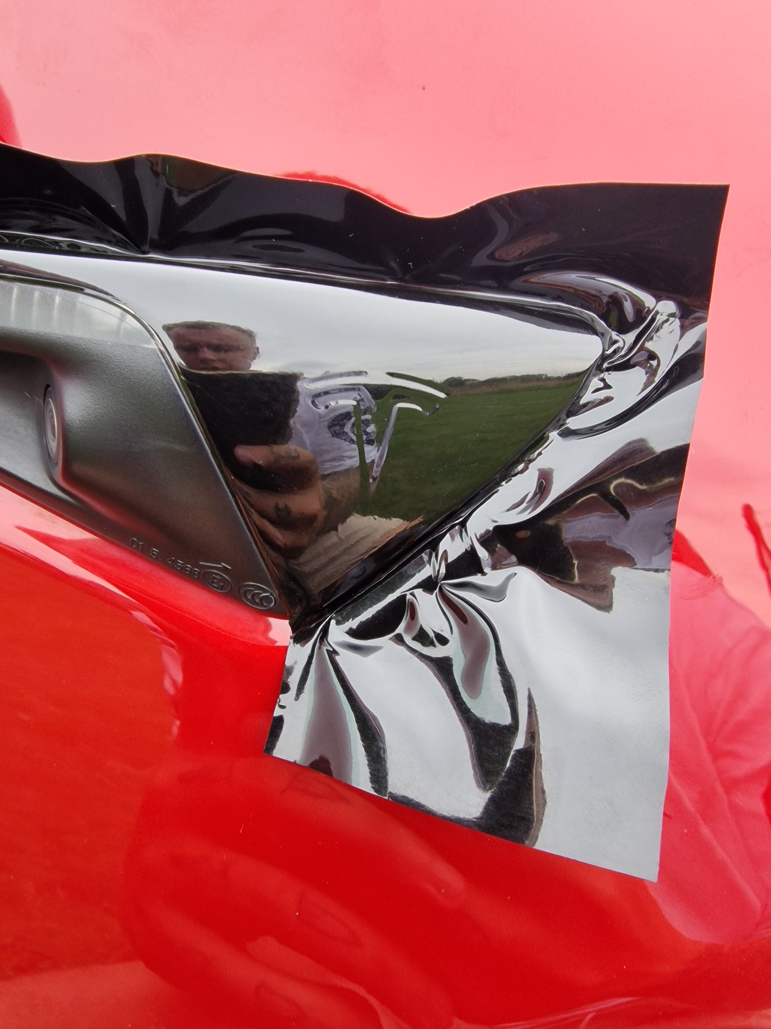 Fragment czerwonego samochodu z naklejoną czarną folią, widoczne odbicie fotografa i otoczenia w połyskującej powierzchni.