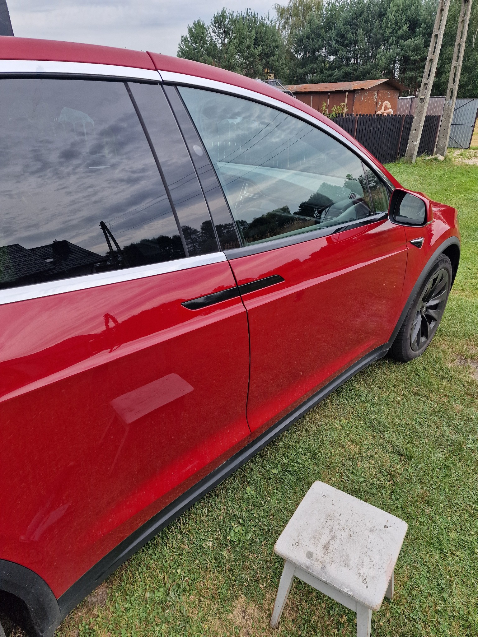 Czerwony samochód elektryczny Tesla Model X po umyciu, z widocznymi smugami wody na lakierze, stojący na trawie obok małego, drewnianego stołka.