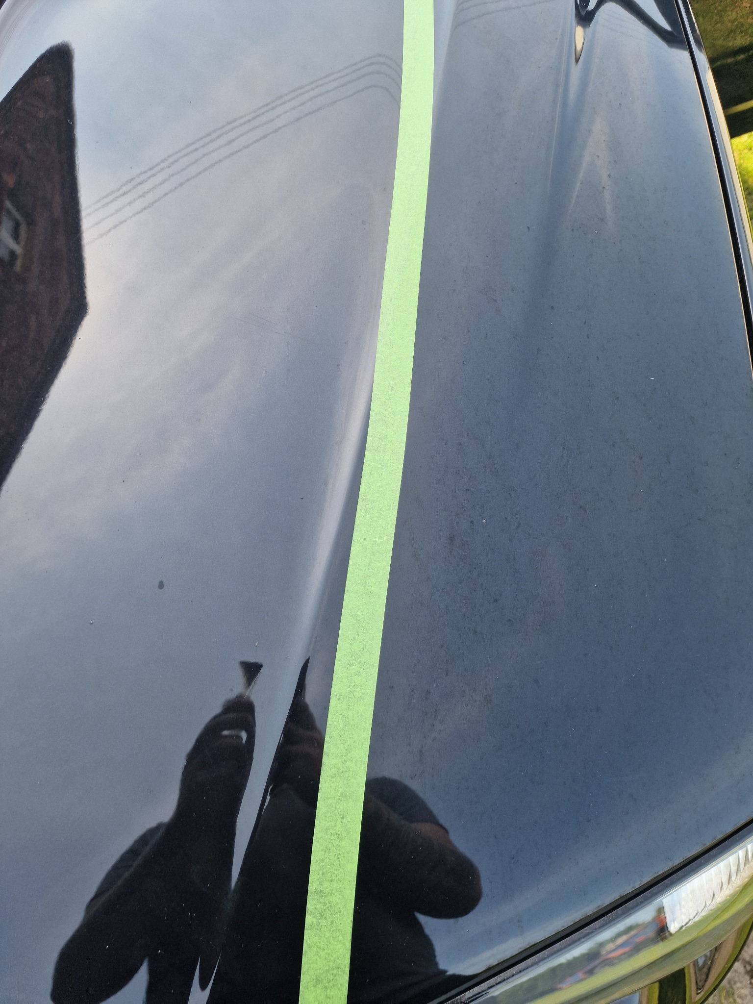 Część maski czarnego samochodu z wyraźnym podziałem: po lewej stronie po polerowaniu, po prawej stronie przed, z zieloną taśmą maskującą oddzielającą obszary.