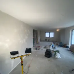 Wnętrze w trakcie prac wykończeniowych, ściany po nałożeniu gładzi, widoczne narzędzia, reflektory oświetleniowe i materiały budowlane, okna zabezpieczone folią.