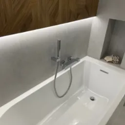 Nowoczesna, biała wanna z chromowaną baterią i deszczownicą, otoczona szarą płytką z podświetleniem LED i dekoracyjną drewnianą mozaiką na ścianie. Wnęka w ścianie z jasną szmatką. Minimalistyczny...