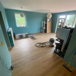 Pokój w trakcie remontu z ułożonymi panelami podłogowymi, niebieskimi ścianami, widocznymi narzędziami, odkurzaczem przemysłowym, oraz stertą paneli w opakowaniach przy ścianie.