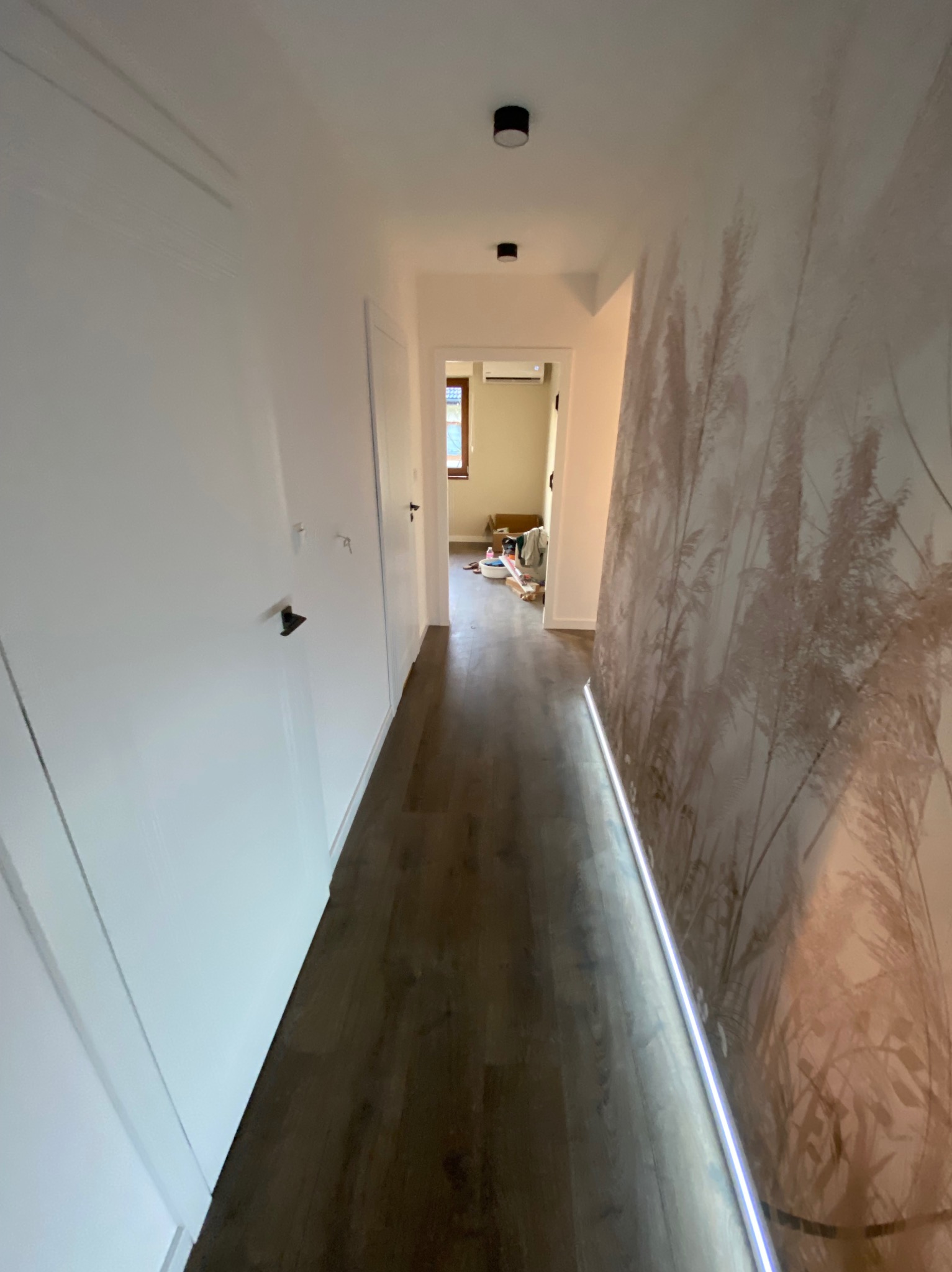 Korytarz w Gdyni po remoncie: ciemne panele podłogowe, białe ściany z szafami w zabudowie oraz dekoracyjna tapeta z motywem roślinnym, oświetlenie punktowe i listwa LED.