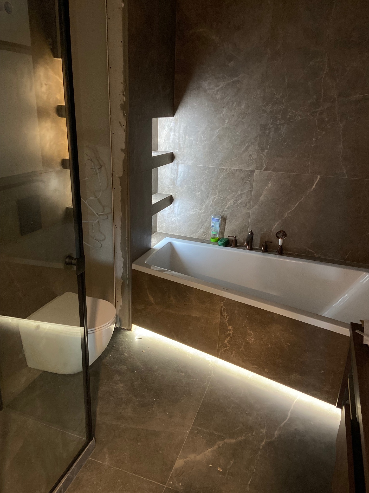 Łazienka w trakcie wykańczania: widoczny fragment kabiny prysznicowej z toaletą, wanna z podświetleniem LED, brązowe płytki imitujące marmur na ścianach i podłodze, fragment regału wnękowego...