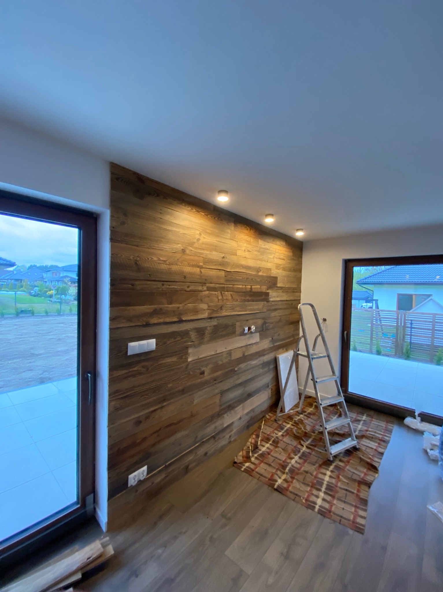 Ściana wykończona panelami drewnianymi z widocznymi gniazdkami elektrycznymi, drabina aluminiowa oparta o ścianę, zabezpieczona podłoga brązową tkaniną w kratę, widoczne okna i drzwi balkonowe...