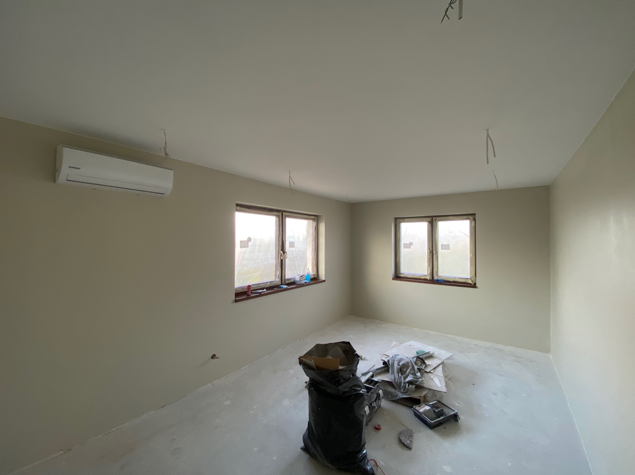 Wnętrze pokoju po malowaniu na odcień beżu, widoczny klimatyzator na ścianie, okna z zabezpieczeniami, ślady po kablach na suficie i worki na śmieci z narzędziami malarskimi.