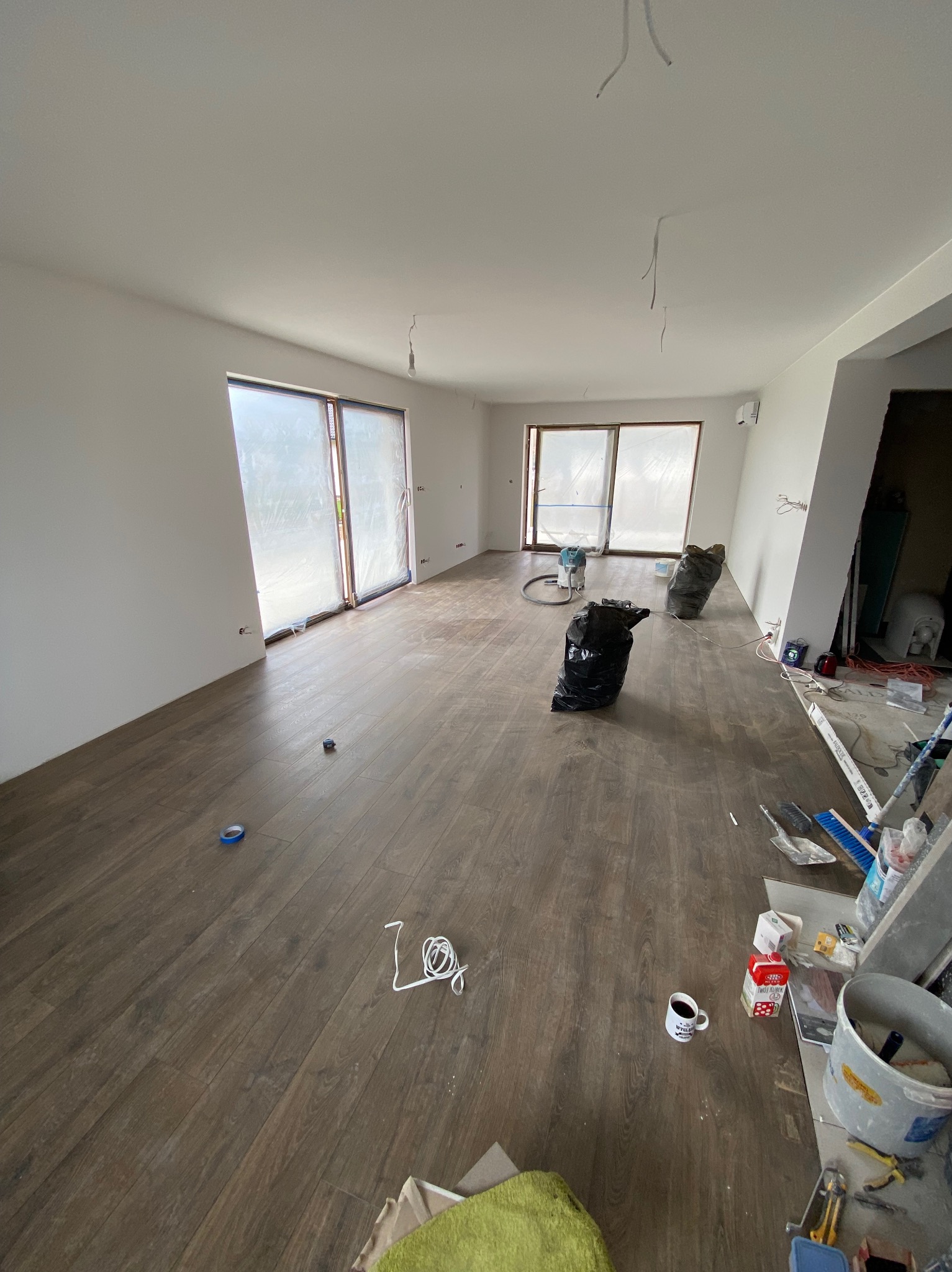 Pokój w trakcie remontu z panelami podłogowymi imitującymi drewno, ściany przygotowane do malowania, okna zabezpieczone folią, widoczne worki na śmieci, narzędzia i kubek z kawą.