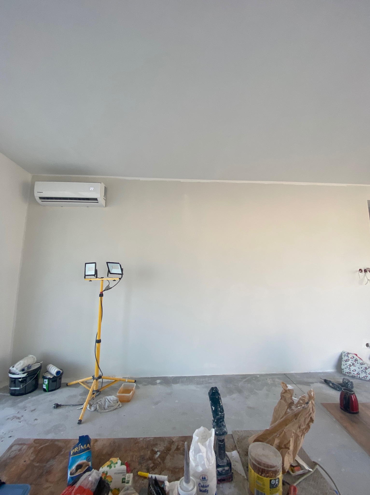 Świeżo pomalowane ściany w pomieszczeniu z zamontowanym klimatyzatorem, widoczne narzędzia malarskie, lampa budowlana i resztki materiałów na podłodze.