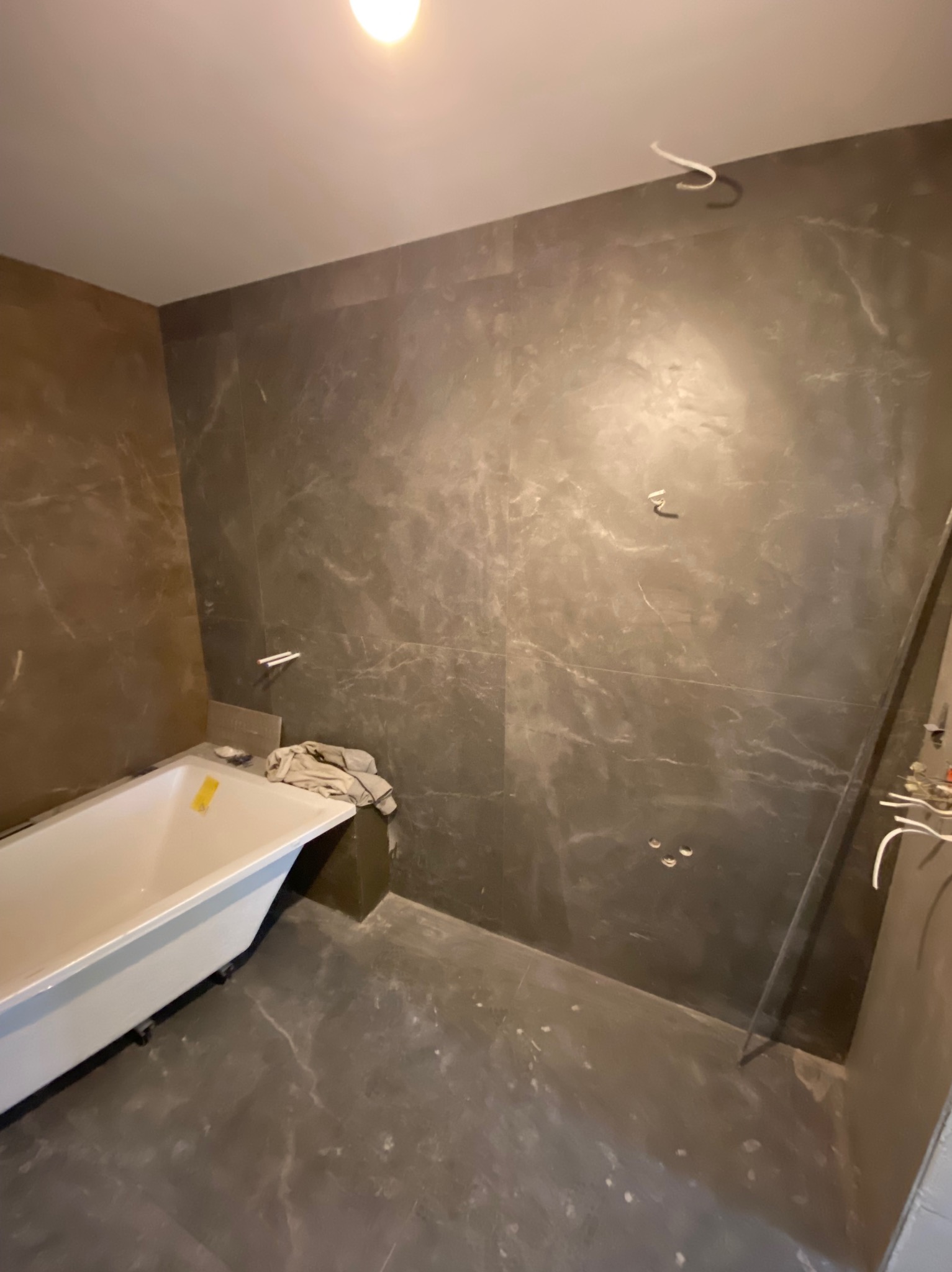 Łazienka w trakcie remontu: biała wanna wolnostojąca na tle ciemnych, marmurkowych płytek ściennych i podłogowych, widoczne wystające kable i elementy instalacji, przygotowania do armatury.