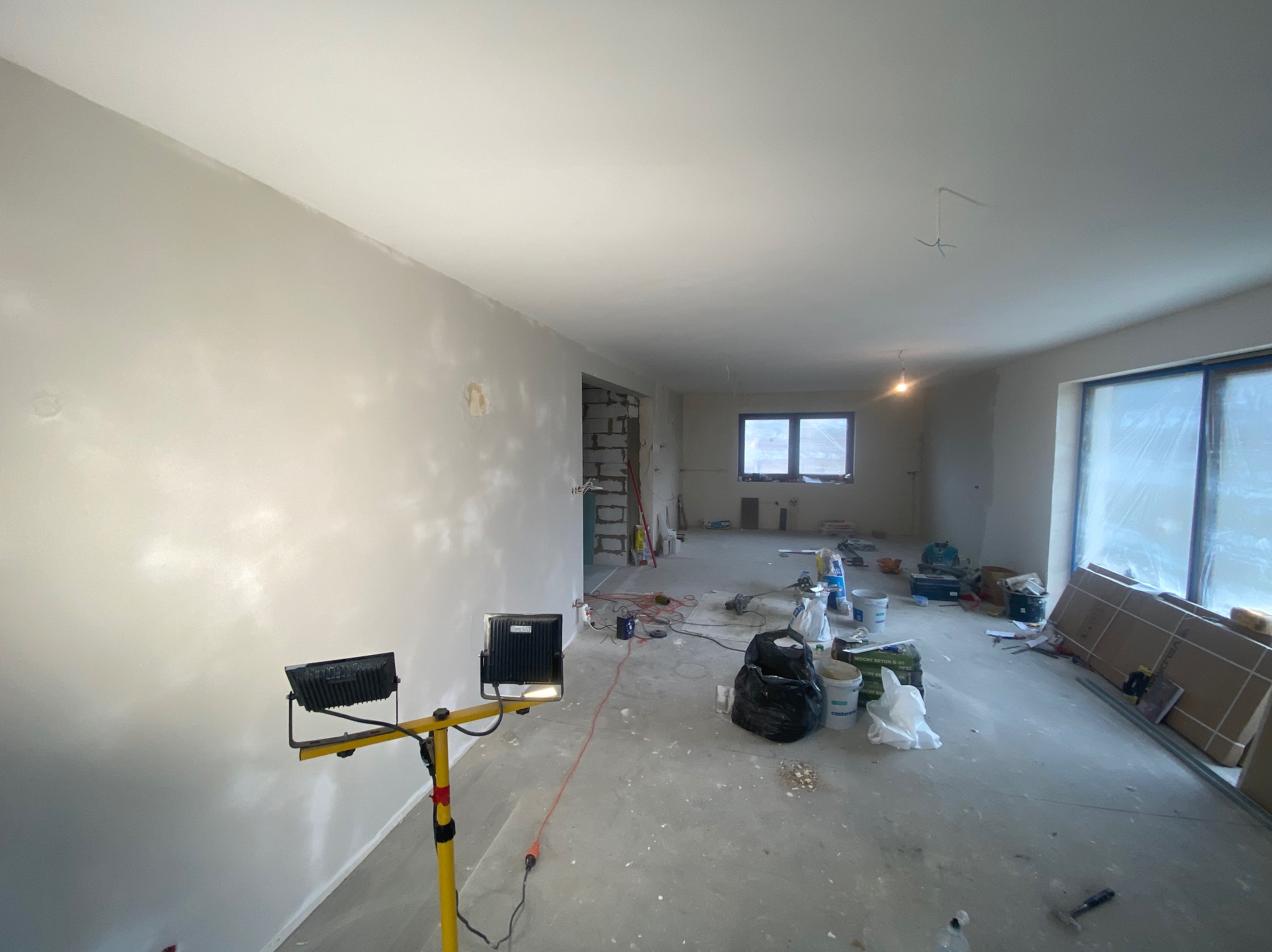Wnętrze w trakcie prac wykończeniowych, ściany po nałożeniu gładzi, widoczne narzędzia, reflektory oświetleniowe i materiały budowlane, okna zabezpieczone folią.