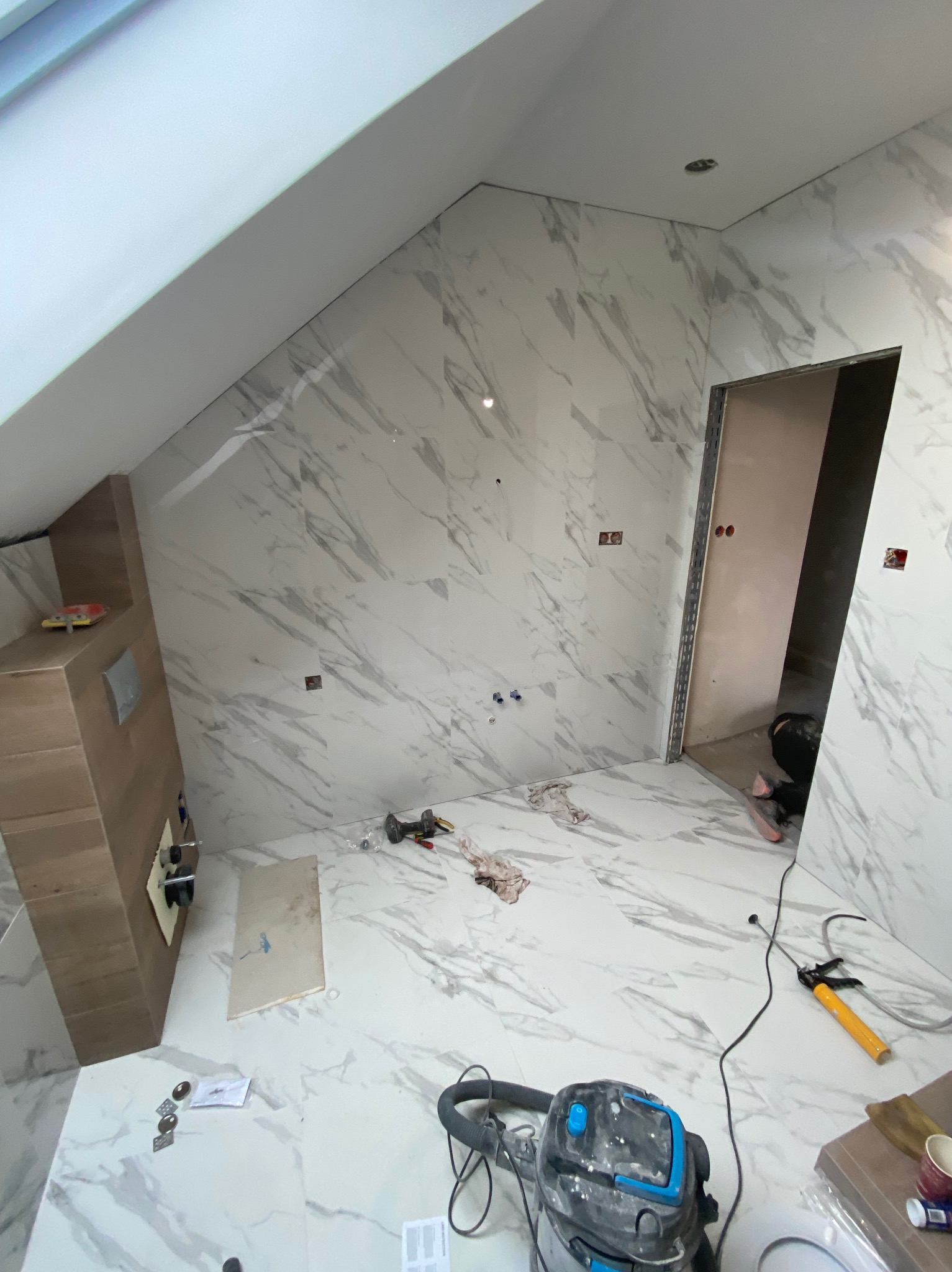 Łazienka w trakcie prac wykończeniowych: marmurowe płytki na ścianach i podłodze, widoczny fragment zabudowy podtynkowego WC, odkurzacz przemysłowy na podłodze, narzędzia i osoba pracująca w tle...