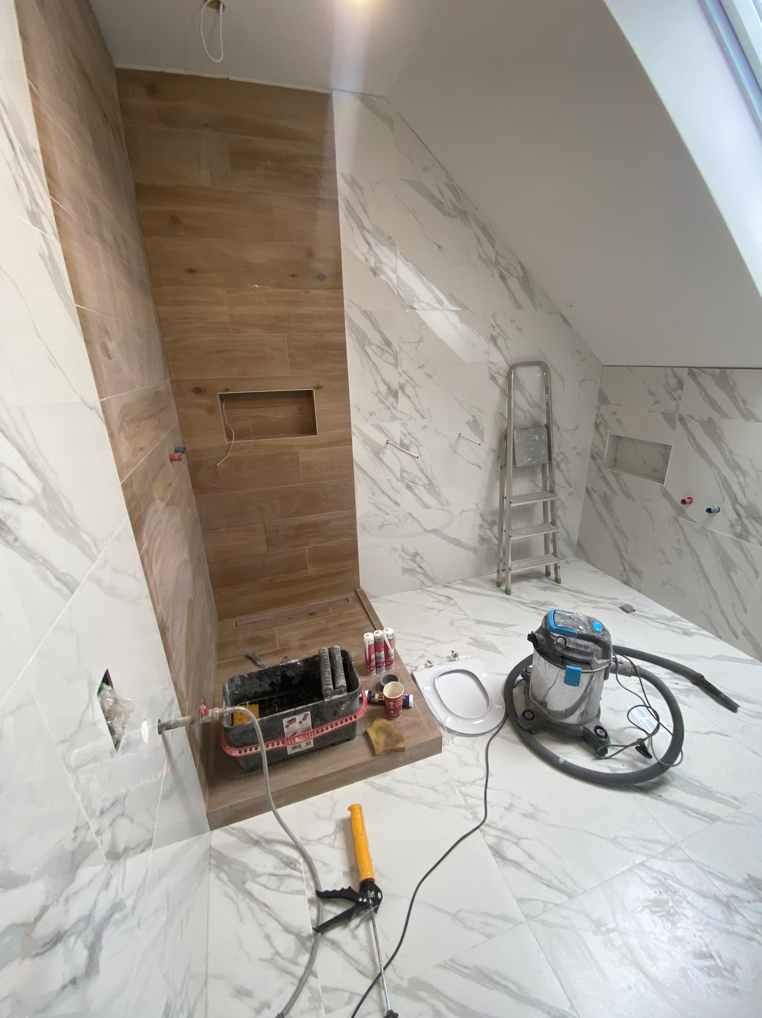 Łazienka w trakcie wykańczania: brązowe płytki imitujące drewno w strefie prysznica, białe marmurowe płytki na ścianach i podłodze, drabina, odkurzacz przemysłowy i narzędzia.