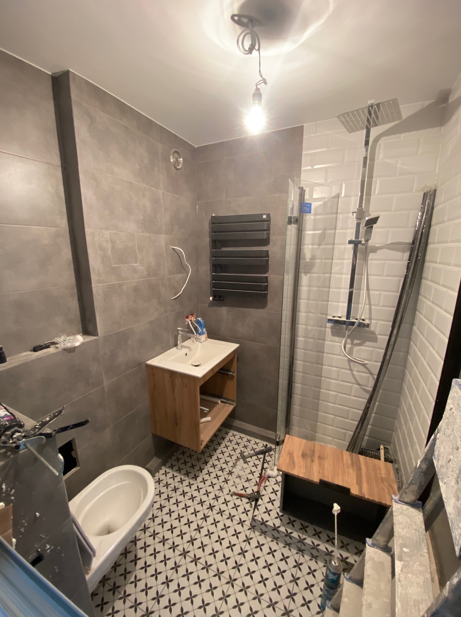 Łazienka w trakcie remontu: szare płytki ścienne, wzorzysta podłoga, zamontowana umywalka z szafką, grzejnik drabinkowy, kabina prysznicowa w trakcie montażu, widoczne narzędzia i prowizoryczne...