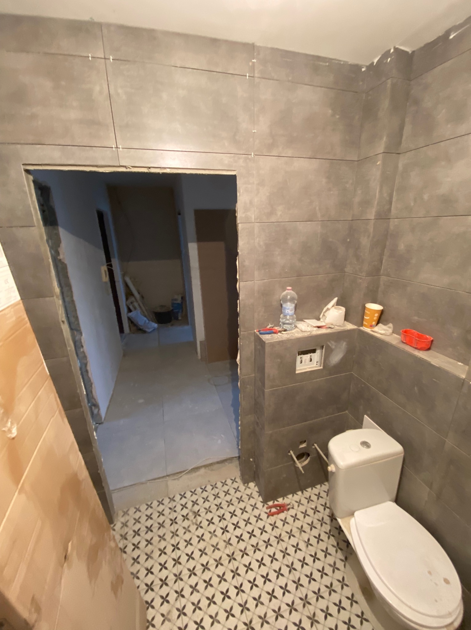 Łazienka w trakcie remontu: szare płytki ścienne ułożone z zachowaniem fug, wzorzysta podłoga, wisząca muszla klozetowa, widoczne narzędzia i materiały budowlane, otwarte przejście do innego...