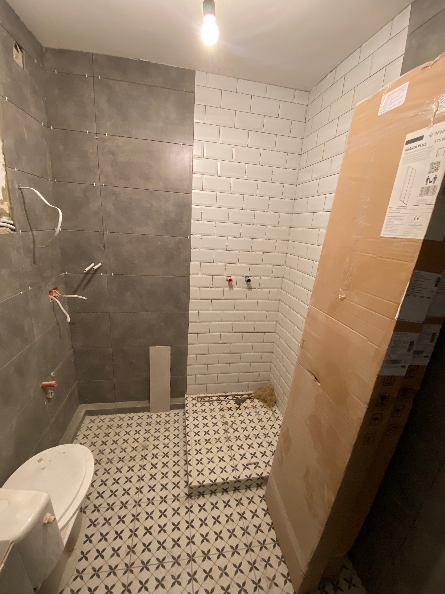 Łazienka w trakcie remontu: częściowo ułożone szare i białe płytki, widoczne instalacje hydrauliczne, karton z nową kabiną prysznicową oparty o ścianę, podłoga z wzorzystych płytek.