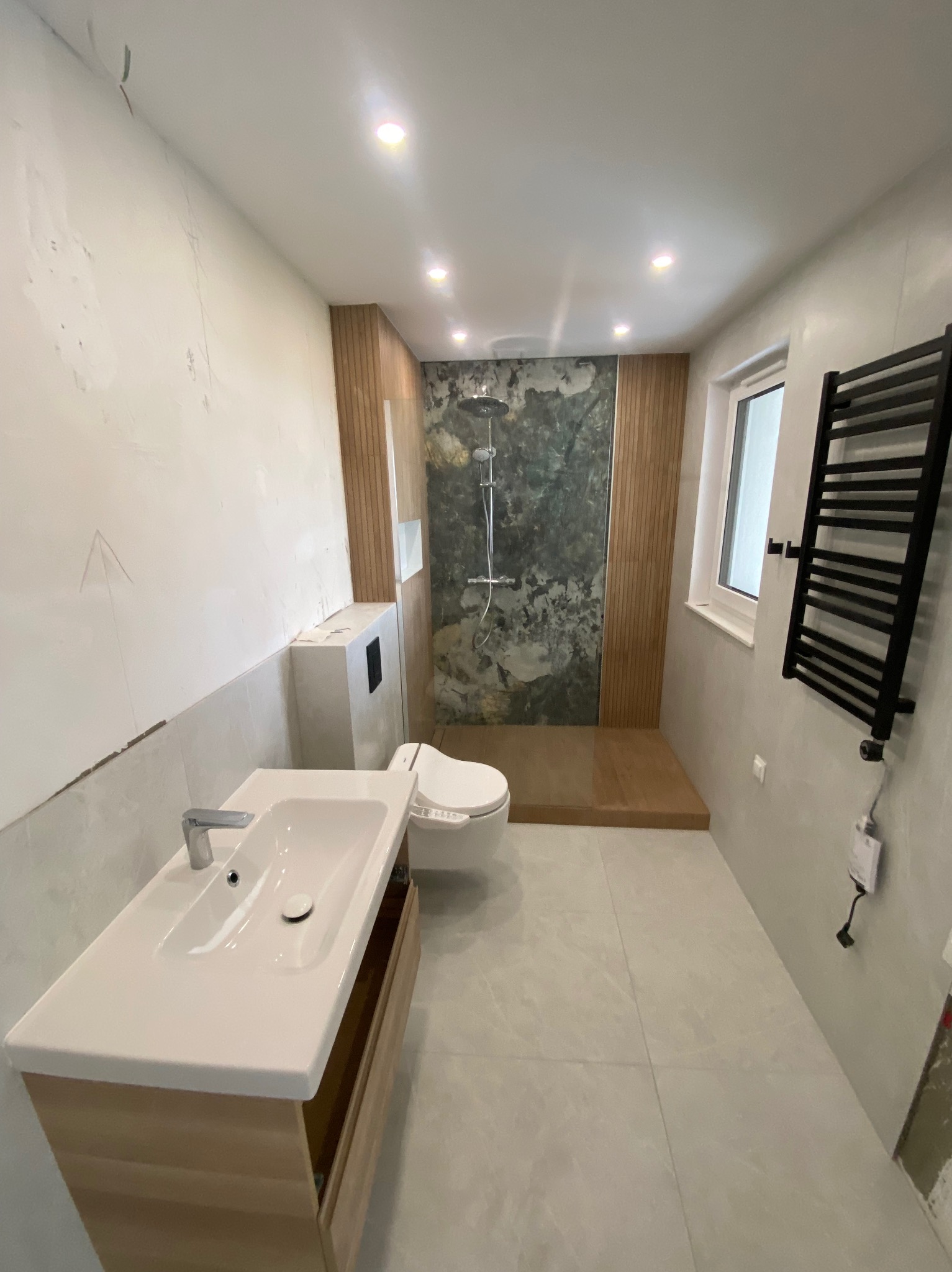 Wnętrze łazienki w trakcie prac wykończeniowych, widoczny fragment ściany bez wykończenia, umywalka z szafką, toaleta, kabina prysznicowa z zielonym panelem dekoracyjnym i grzejnik drabinkowy.