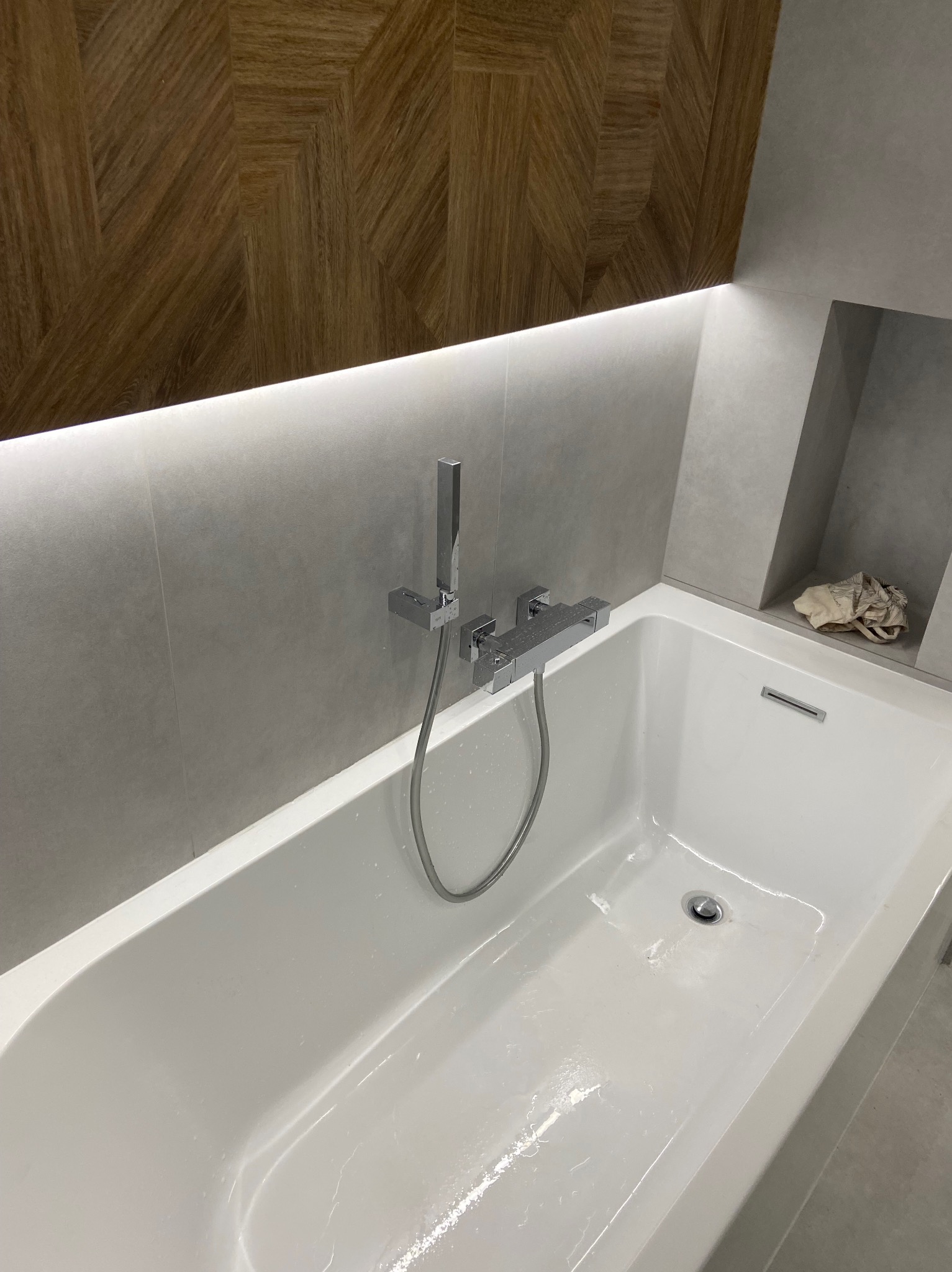 Nowoczesna, biała wanna z chromowaną baterią i deszczownicą, otoczona szarą płytką z podświetleniem LED i dekoracyjną drewnianą mozaiką na ścianie. Wnęka w ścianie z jasną szmatką. Minimalistyczny...