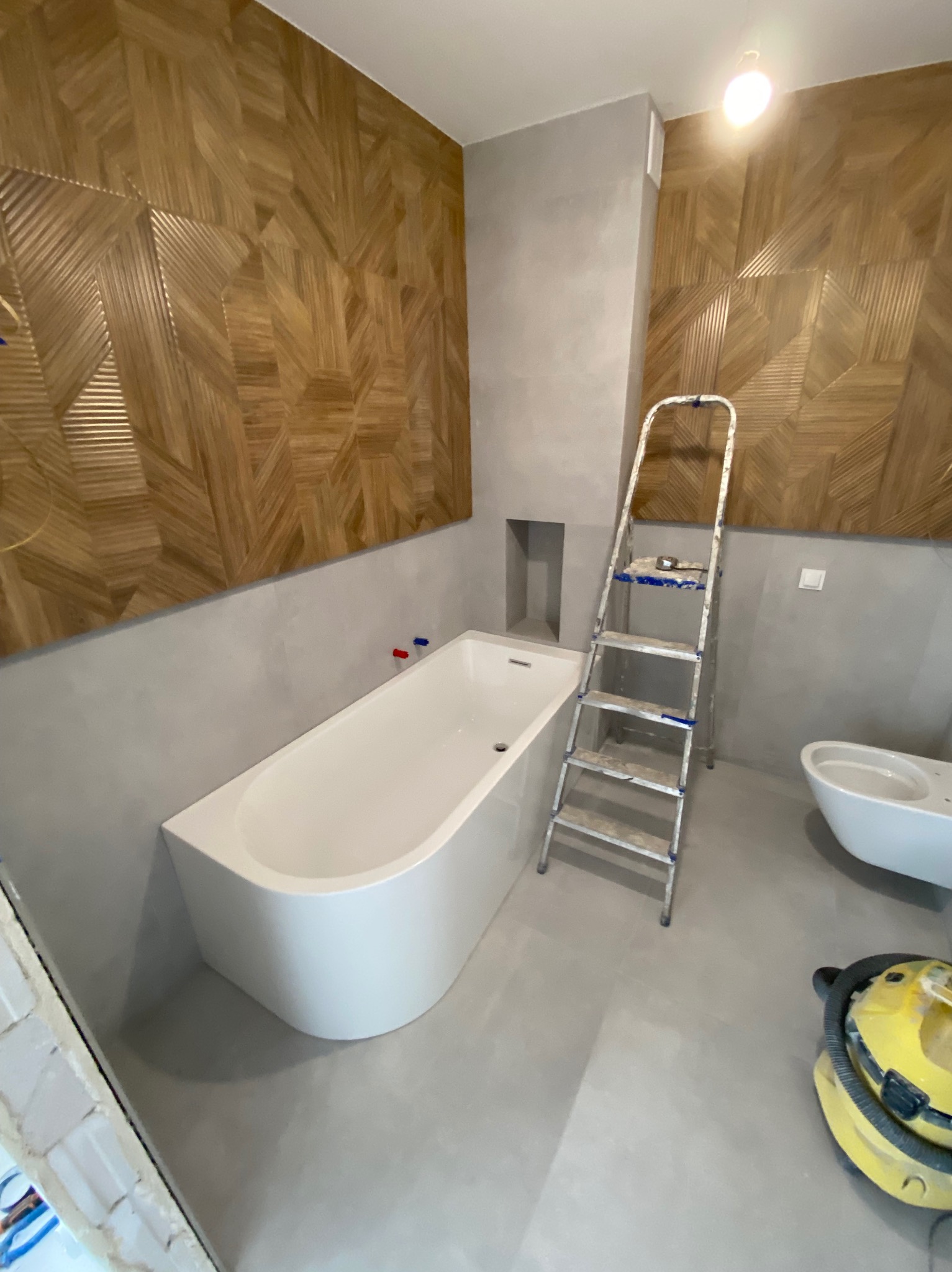 Łazienka w trakcie wykańczania: biała wanna, drewniana mozaika na ścianie, szare ściany, drabina malarska, częściowo zamontowana toaleta, odkurzacz budowlany na podłodze.