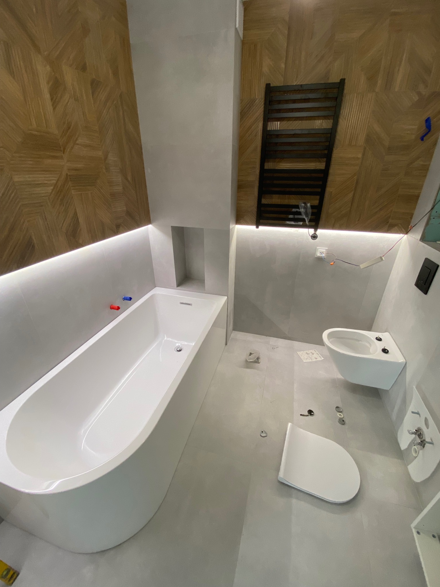 Łazienka w trakcie wykańczania: biała wanna z podświetleniem LED, wisząca toaleta i bidet, grzejnik drabinkowy, drewniana ściana z geometrycznym wzorem, widoczne elementy instalacji i narzędzia.