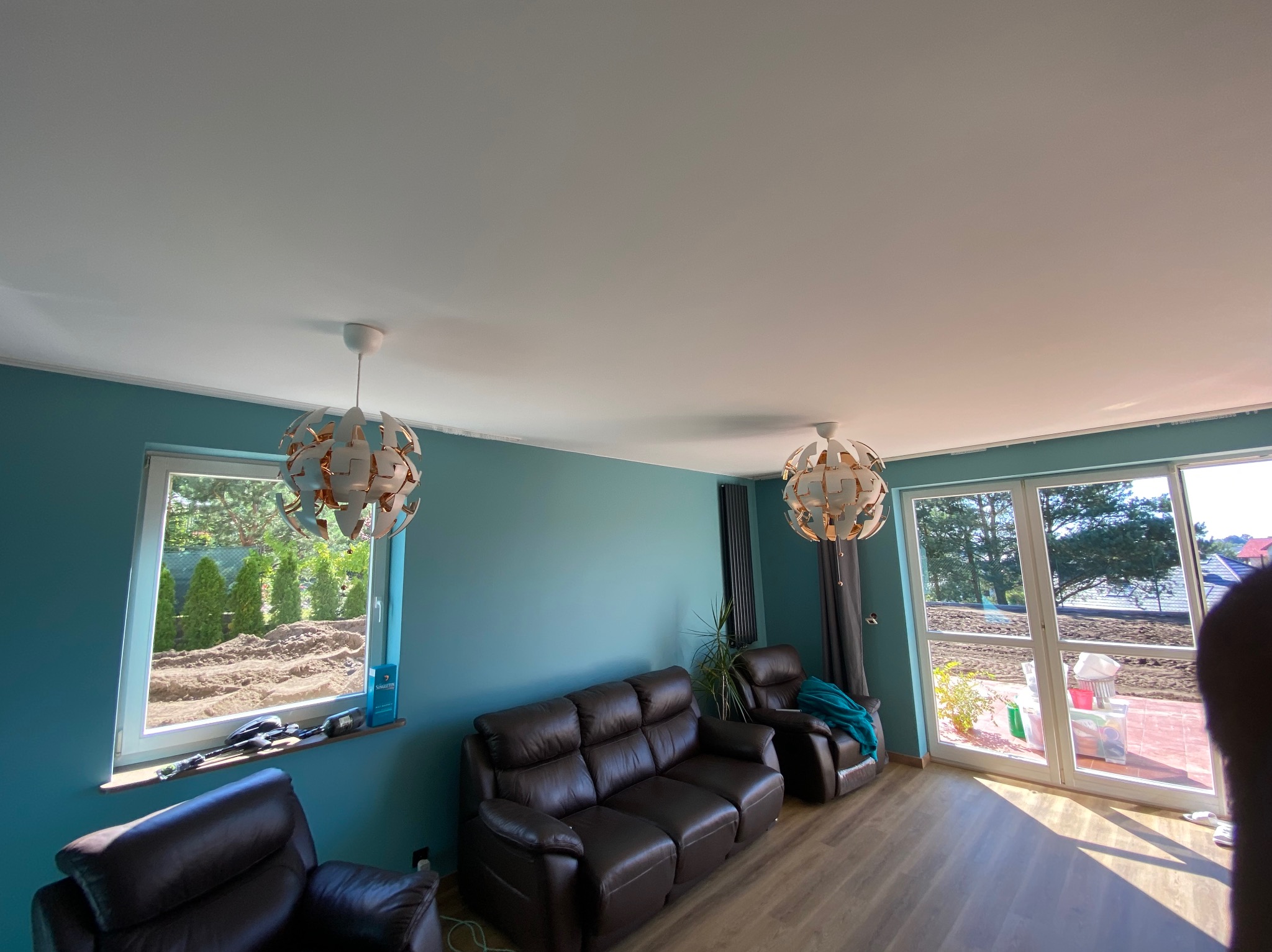 Turkusowa ściana w salonie z brązową skórzaną kanapą i dwoma lampami wiszącymi o geometrycznym designie, widok na ogród przez okno.