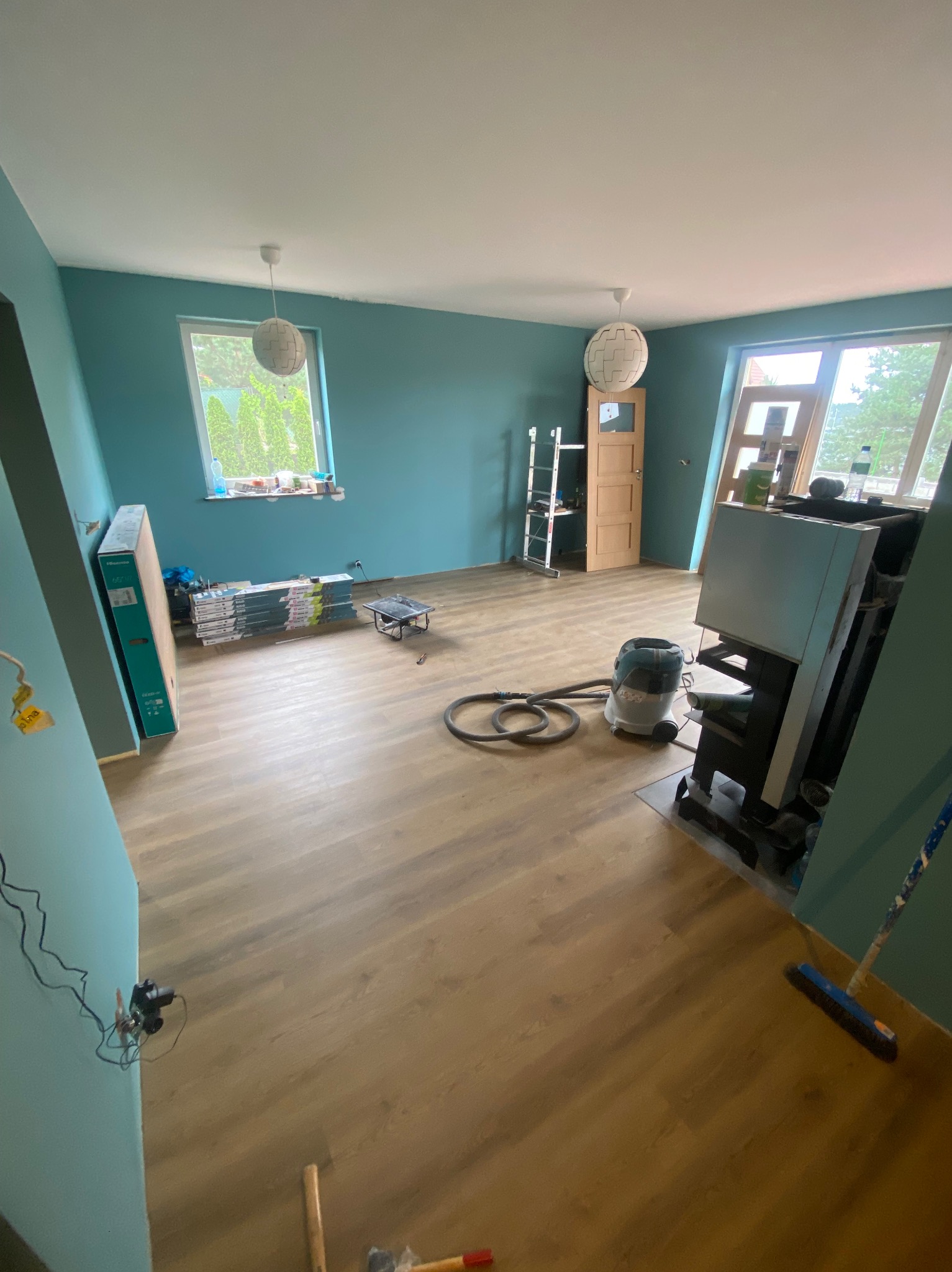 Pokój w trakcie remontu z ułożonymi panelami podłogowymi, niebieskimi ścianami, widocznymi narzędziami, odkurzaczem przemysłowym, oraz stertą paneli w opakowaniach przy ścianie.