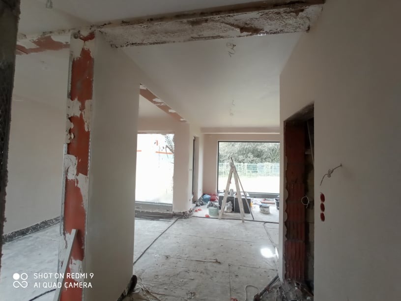 Wnętrze w trakcie tynkowania: surowe filary z widoczną cegłą i resztkami tynku, świeżo otynkowane ściany i sufit, drabina w tle przy oknie.