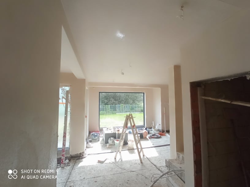 Wnętrze w trakcie prac wykończeniowych, świeżo nałożone gładzie na ścianach i suficie, widoczne narzędzia i drabina, duże okno z widokiem na zieleń w tle.