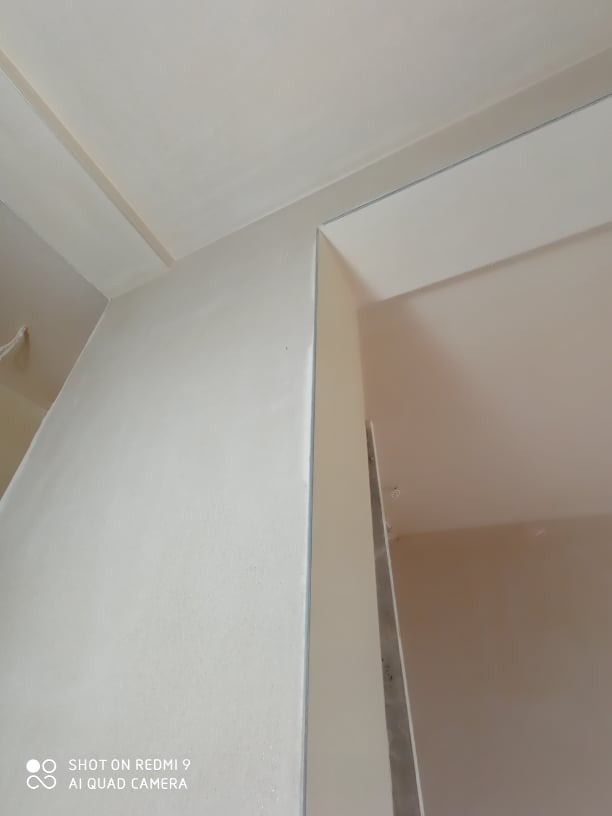 Świeżo otynkowana ściana i sufit w jasnym odcieniu, widoczne subtelne nierówności faktury, kąt ujęcia od dołu, z widocznym fragmentem otworu drzwiowego w tle.