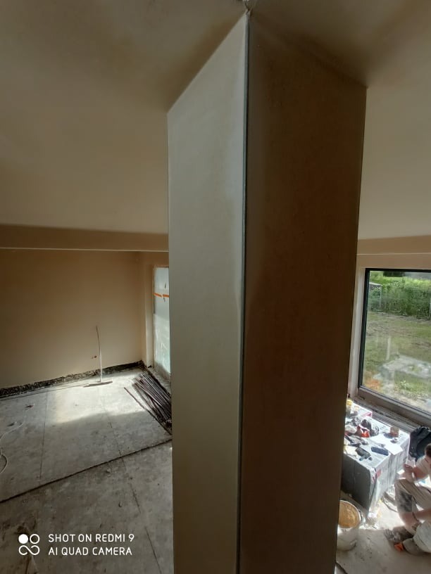 Wnętrze w trakcie prac wykończeniowych; kolumna po nałożeniu świeżej, gładkiej warstwy tynku, widoczne narzędzia i wiadro z zaprawą w pobliżu okna.