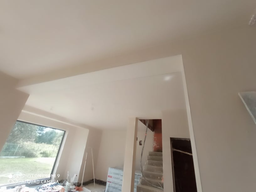 Wnętrze domu w trakcie wykańczania, świeżo położone gładzie na ścianach i suficie, widoczne okno z widokiem na zieleń i fragment schodów.