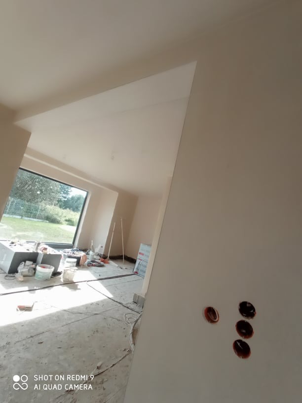 Świeżo otynkowane ściany wewnętrzne w jasnym pomieszczeniu z dużym oknem, widoczne puszki elektryczne bez osprzętu i sprzęt do nakładania tynków w tle.