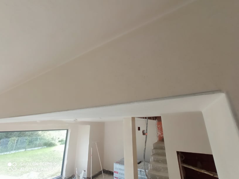 Gładko wykończony sufit pod skosem po nałożeniu gładzi, widoczne przejście między sufitem a ścianą, okno z widokiem na zieleń w tle.