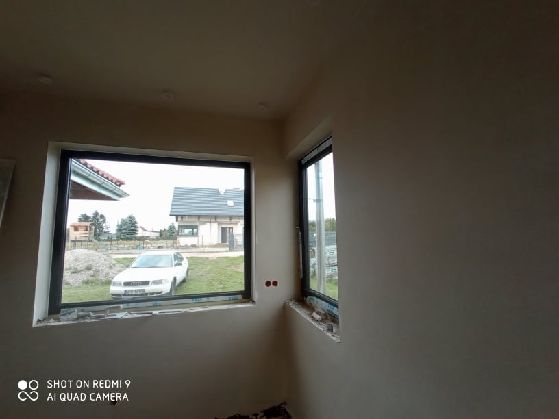 Wnętrze w trakcie prac wykończeniowych, ściany pokryte świeżym tynkiem o jasnym odcieniu, widoczne dwa okna z czarnymi ramami, przez jedno z nich widać biały samochód i dom w tle.