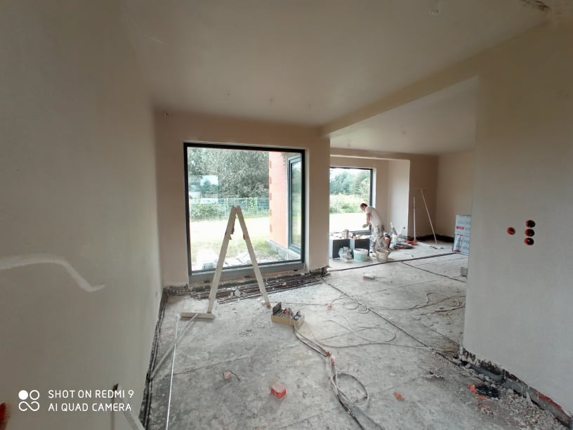 Wnętrze w trakcie prac wykończeniowych: świeżo nałożony tynk na ścianach, drabina oparta o okno, widoczny pracownik w oddali przygotowujący maszynę do tynkowania, kable na podłodze.