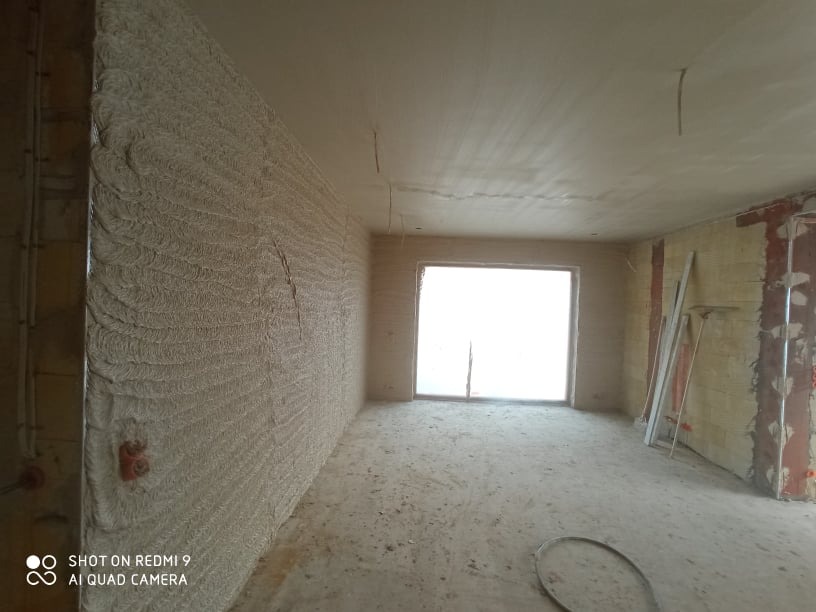 Świeżo nałożony tynk na ścianie wewnętrznej, widoczne ślady po narzędziach, surowy stan wykończenia, okno w tle z jasnym światłem dziennym.