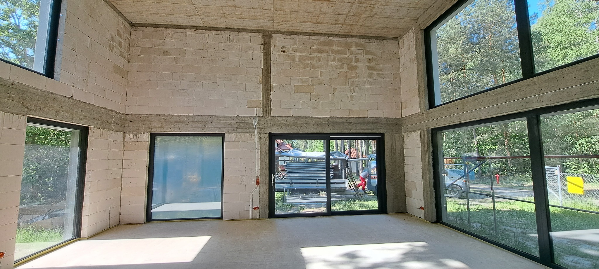 Surowe wnętrze domu w trakcie budowy z widocznymi ścianami z betonu komórkowego, betonowymi nadprożami i dużymi oknami z czarnymi ramami wychodzącymi na las. Na zewnątrz widoczny sprzęt budowlany...