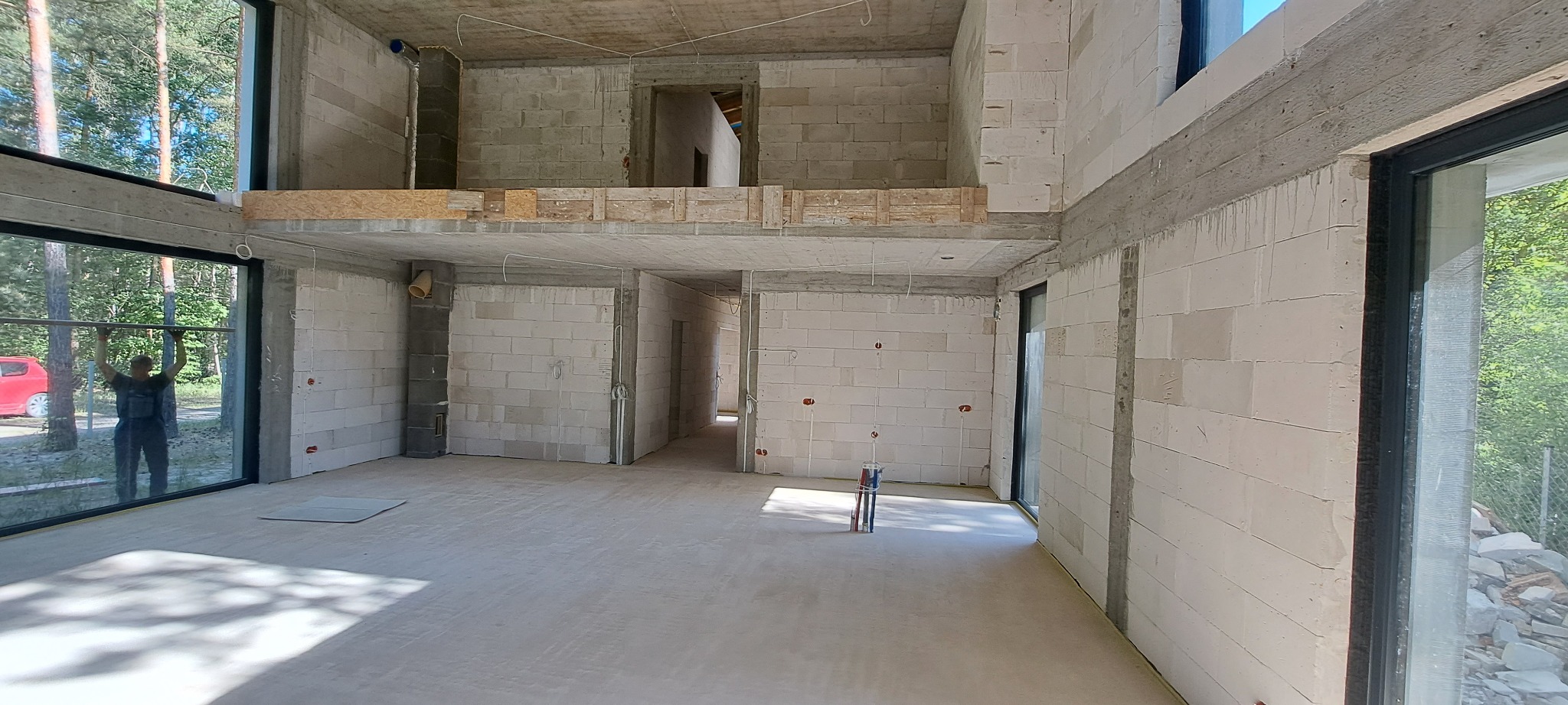 Wnętrze surowego domu w trakcie budowy z widocznymi ścianami z bloczków, wylewką podłogową i instalacjami elektrycznymi; pracownik montuje okno.