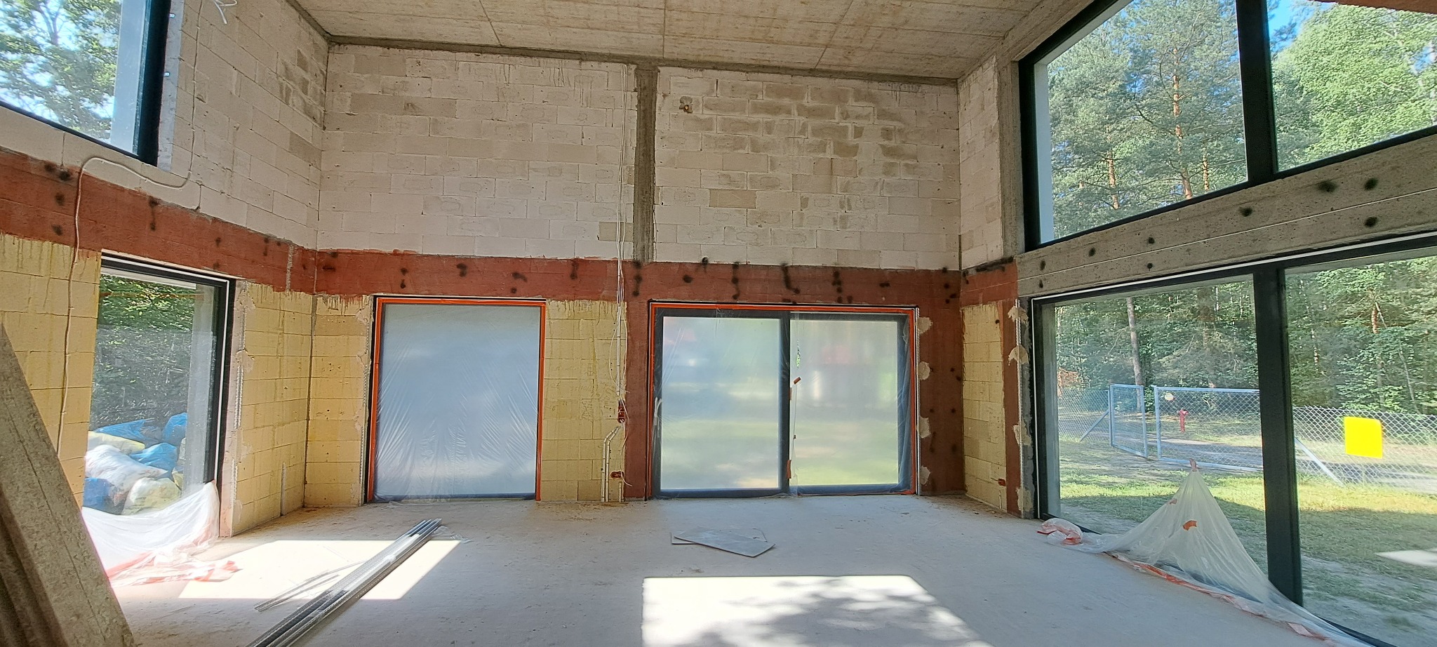 Wnętrze surowego domu w trakcie prac tynkarskich: ściany z bloczków, częściowo ocieplone, z widocznymi śladami po kołkach, okna zabezpieczone folią, na podłodze resztki materiałów budowlanych...