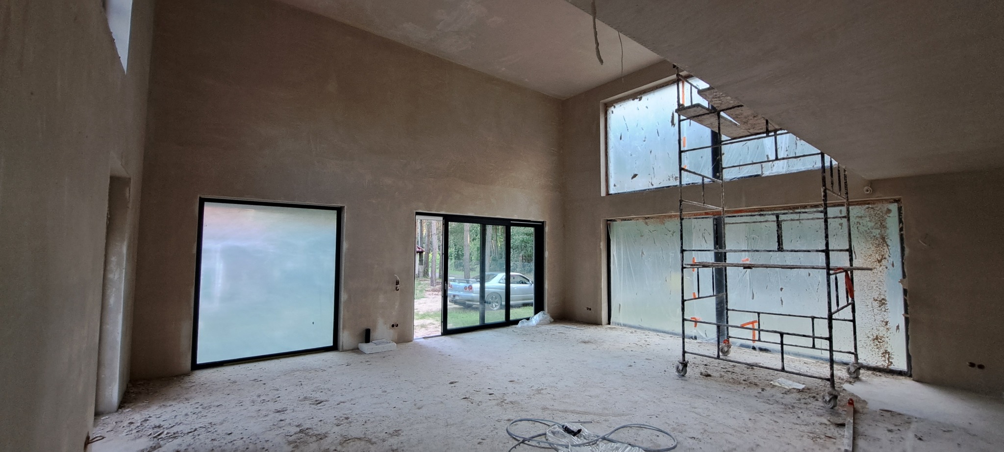 Surowe wnętrze domu w trakcie prac wykończeniowych, ściany pokryte świeżym tynkiem, widoczne rusztowanie przy dużym oknie, okna zabezpieczone folią malarską.