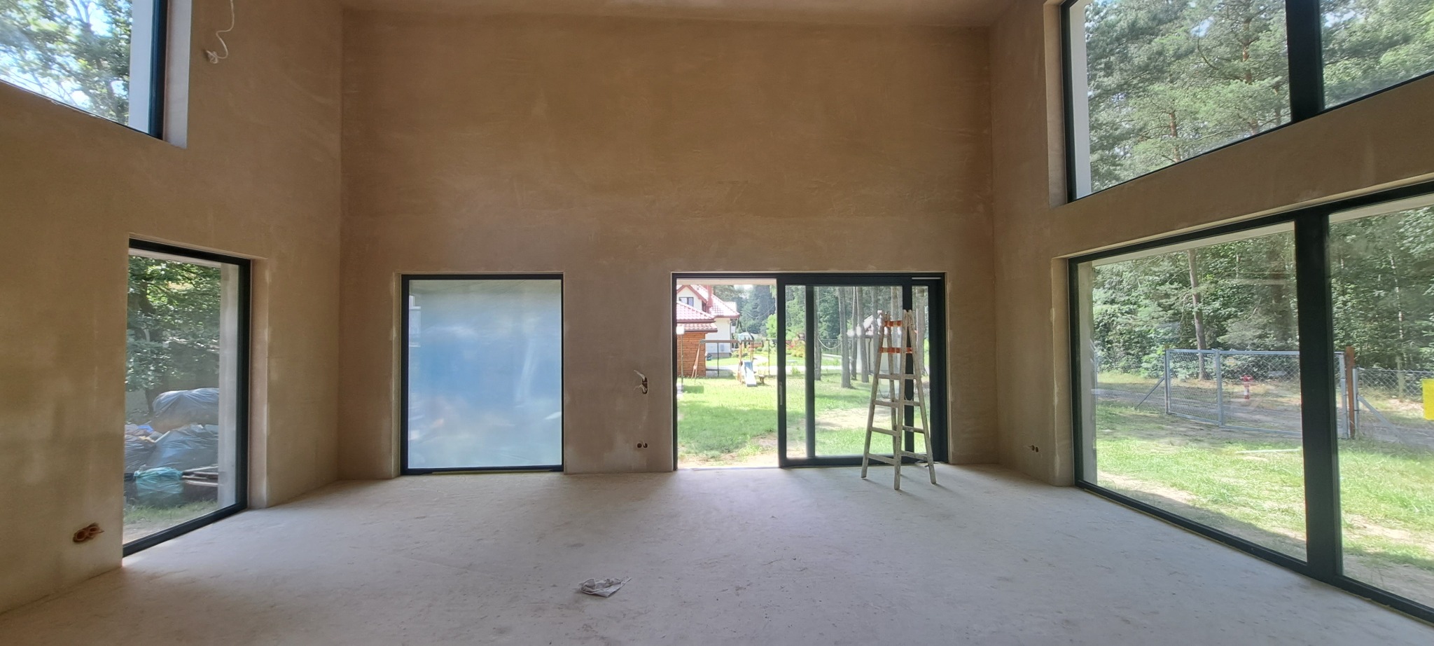 Surowe, jasne wnętrze domu po nałożeniu tynku, z widoczną drabiną, dużymi oknami z widokiem na zieleń i przesuwne drzwi balkonowe.