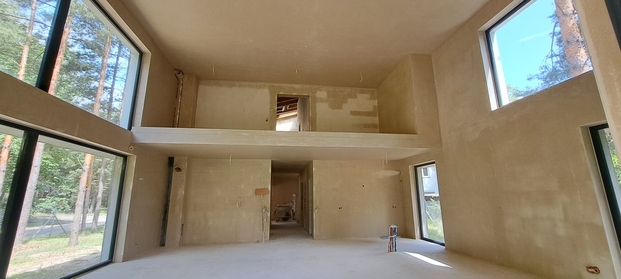 Surowe wnętrze domu z antresolą po nałożeniu tynków, widoczne duże okna z widokiem na las, przygotowane pod dalsze prace wykończeniowe.