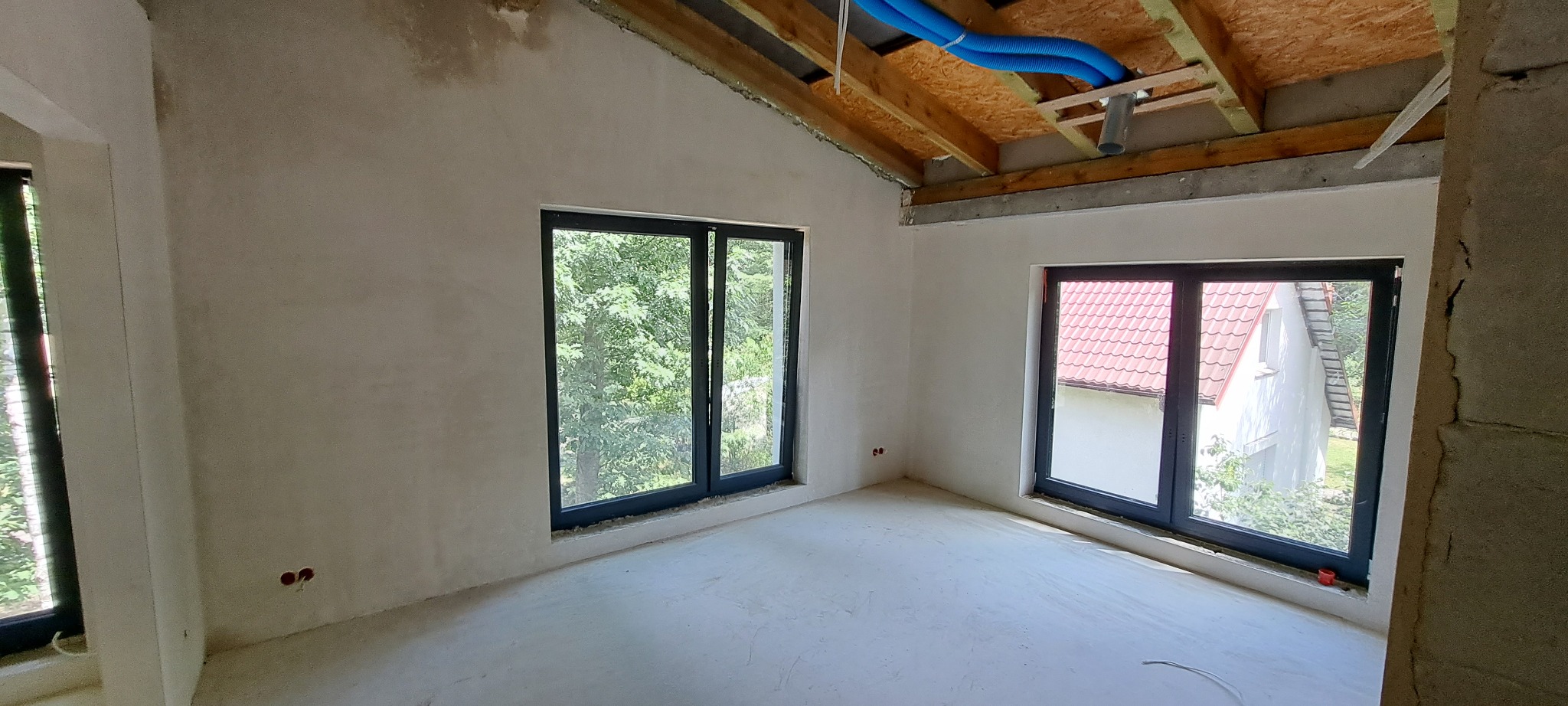 Surowe wnętrze po nałożeniu tynku, widoczne dwa okna z widokiem na zieleń i dom z czerwoną dachówką, niebieskie rury wentylacyjne pod drewnianym stropem.