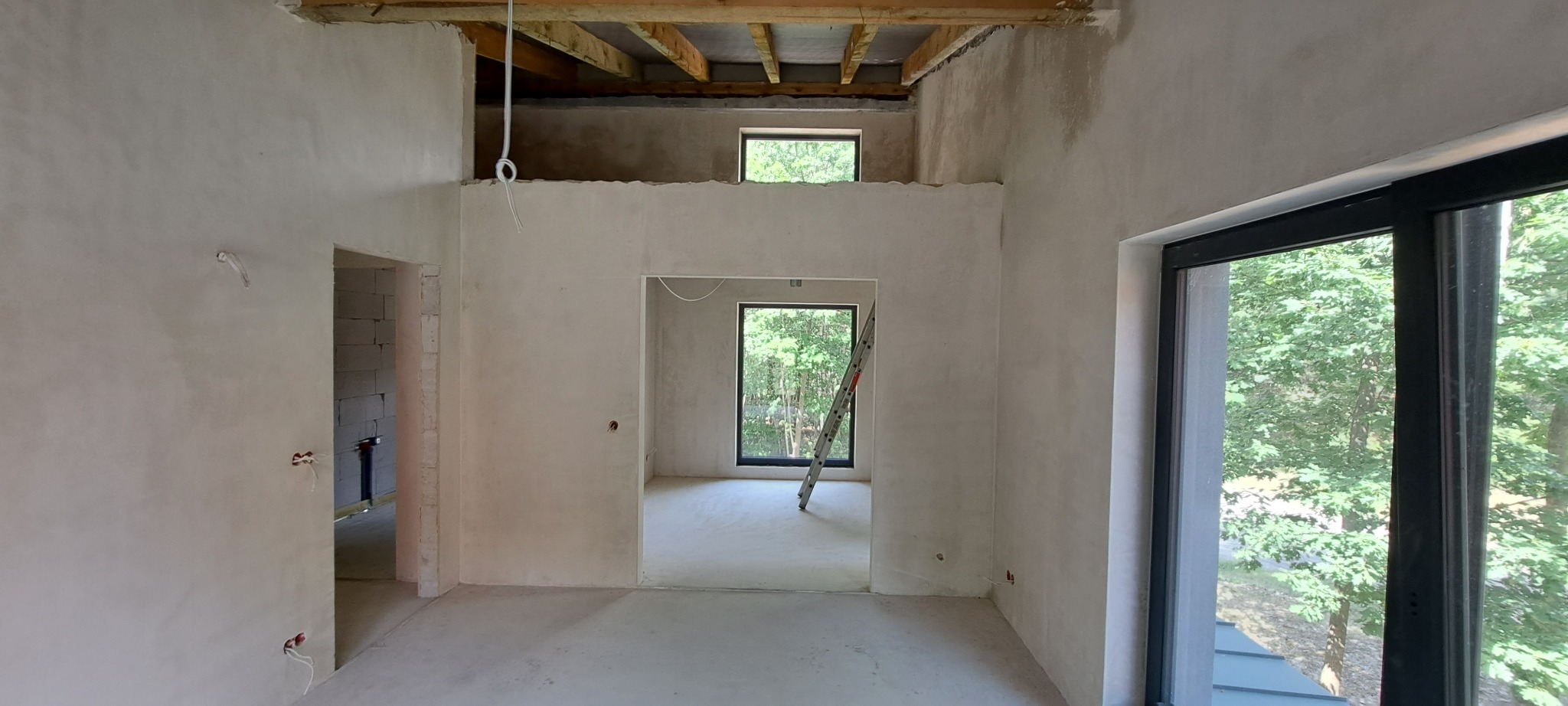 Wnętrze w stanie deweloperskim po nałożeniu gładzi gipsowej na ścianach, widoczne instalacje elektryczne, surowy strop drewniany, okna z widokiem na zieleń i drabina oparta o ścianę.