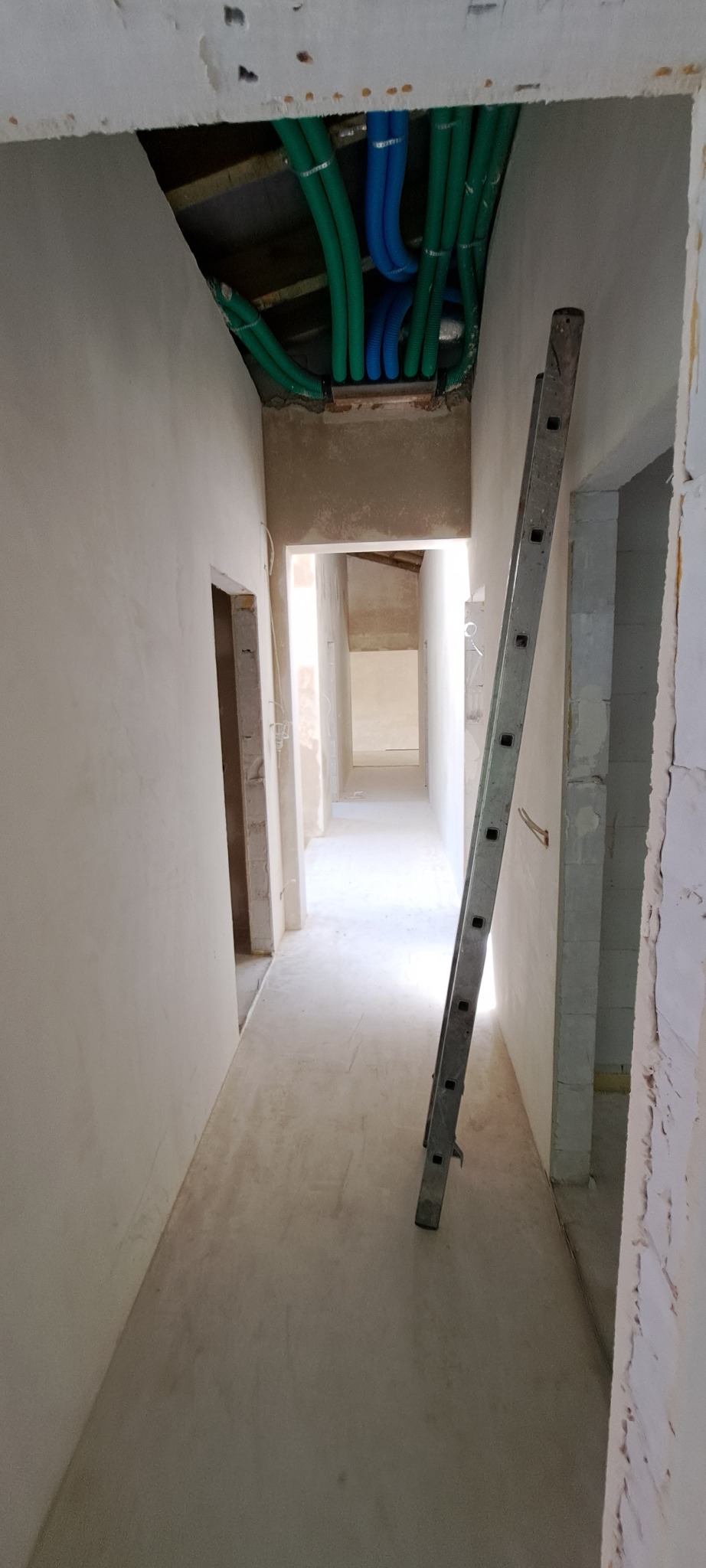 Długi korytarz w stanie surowym z widocznymi instalacjami w stropie i drabiną opartą o ścianę.