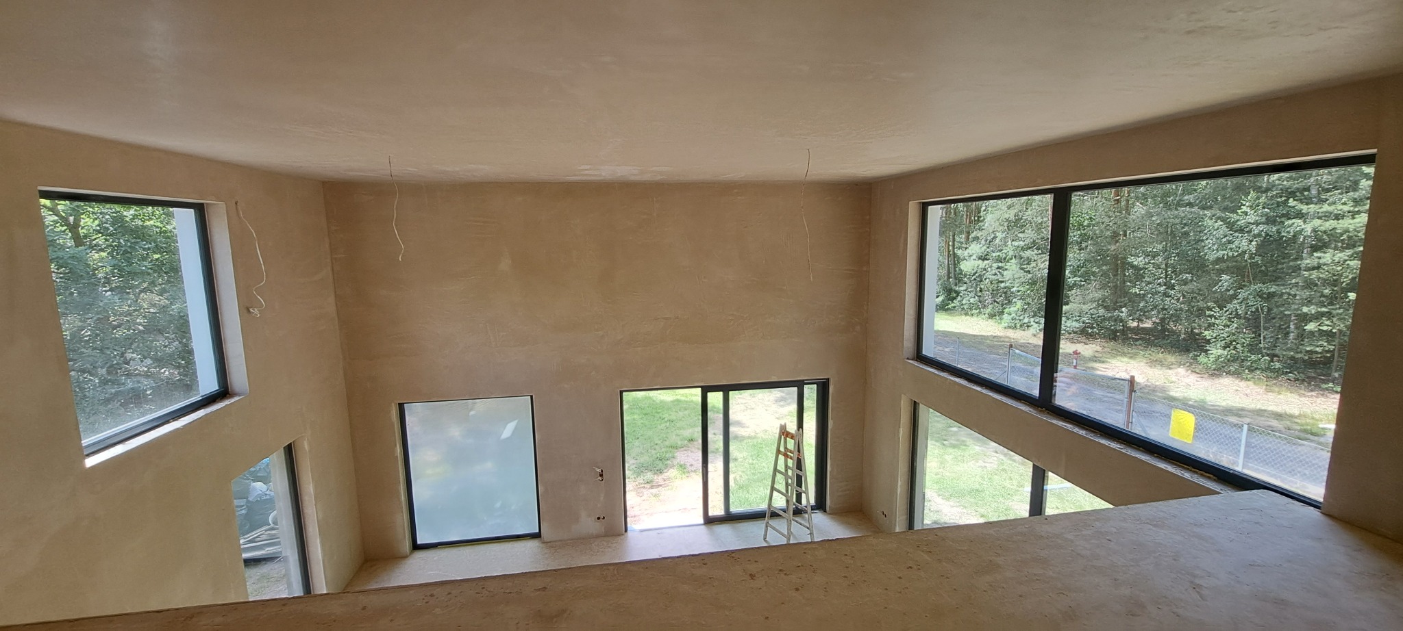 Wnętrze domu po tynkowaniu maszynowym, widok z góry na dwukondygnacyjną przestrzeń z dużymi oknami z czarnymi ramami, widoczna drabina oparta o ścianę, surowy stan wykończenia.