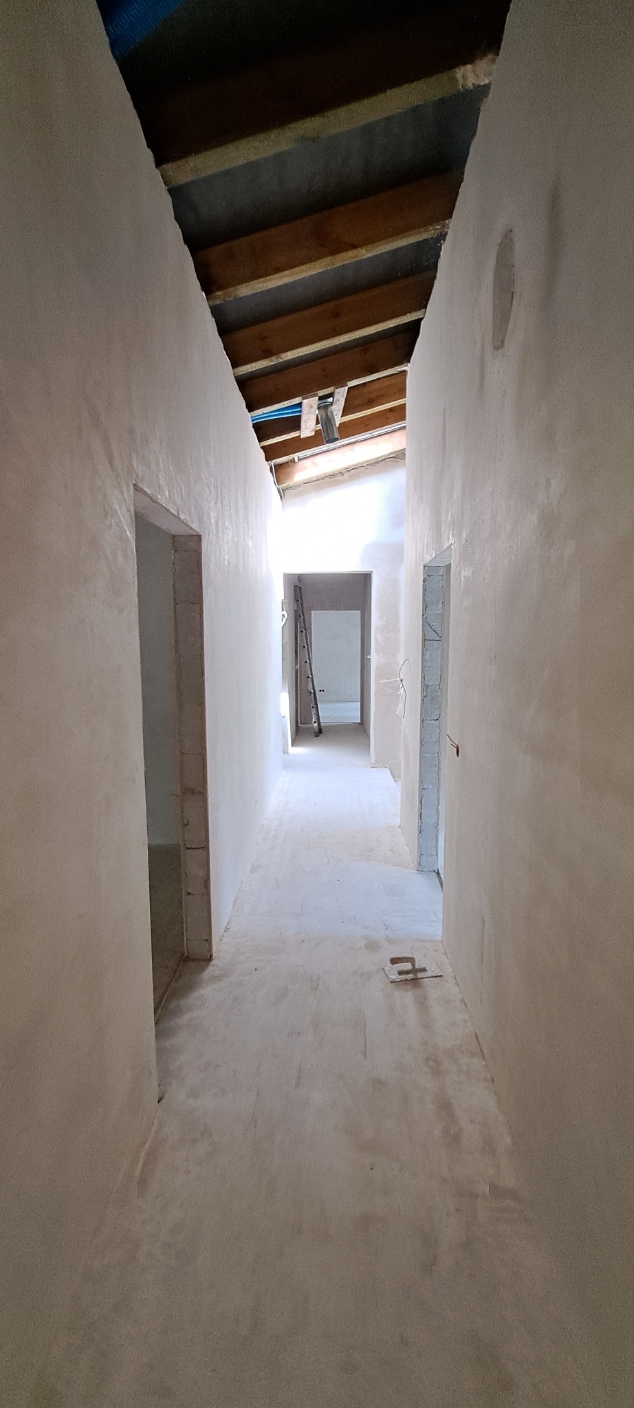 Długi korytarz w trakcie prac wykończeniowych, świeżo położone tynki na ścianach, widoczna drabina oparta o ścianę w głębi, surowy strop drewniany i narzędzie na podłodze.