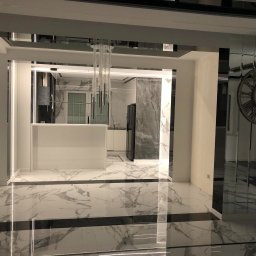 Dawid Żyłka - Nowoczesne wnętrze z marmurową podłogą, widok na białą kuchnię z wyspą, ozdobne lustra i wisząca lampa.