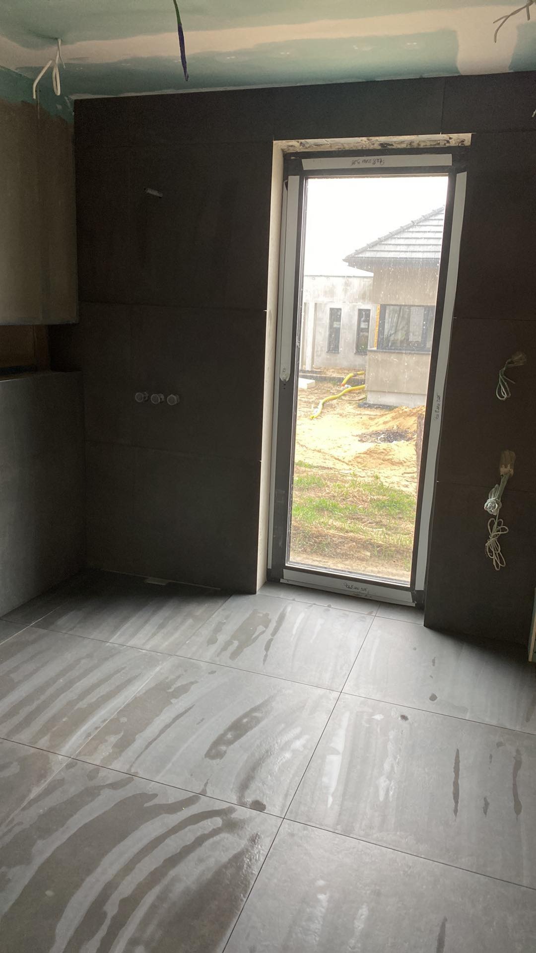 Wnętrze w trakcie prac wykończeniowych: ciemne płytki na ścianach i podłodze, surowy stan sufitu z wystającymi kablami, widok na budowę za oknem, mokra podłoga.