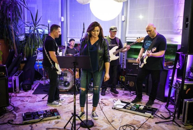 Zespół muzyczny podczas występu w pomieszczeniu z roślinami, wokalistka w skórzanej kurtce i dżinsach śpiewa do mikrofonu, widoczni gitarzyści, perkusista i efekty gitarowe.