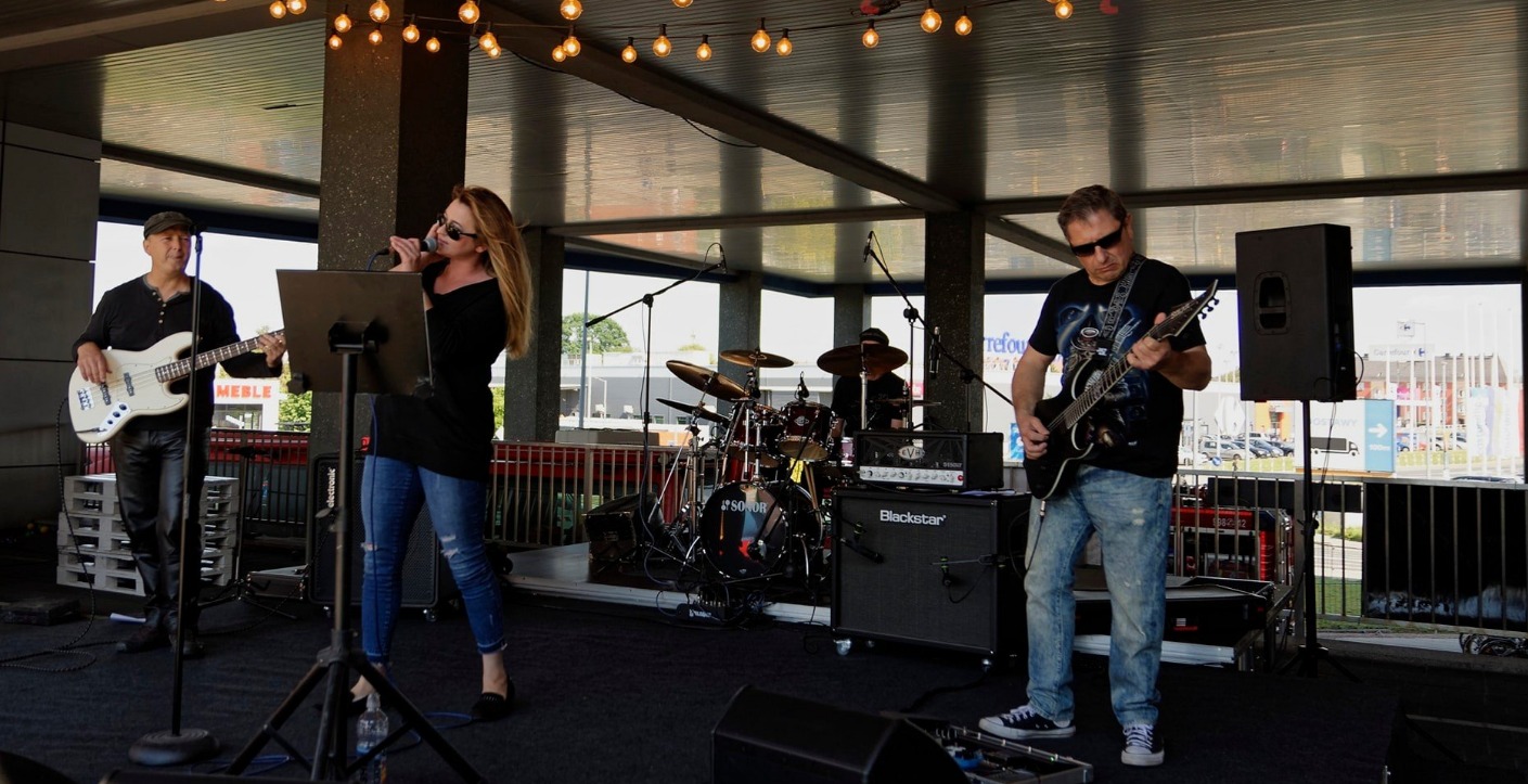 Zespół muzyczny grający koncert na zewnątrz: wokalistka śpiewa do mikrofonu, gitarzysta gra na gitarze elektrycznej, basista gra na basie, a perkusista gra na perkusji. Scena z oświetleniem...